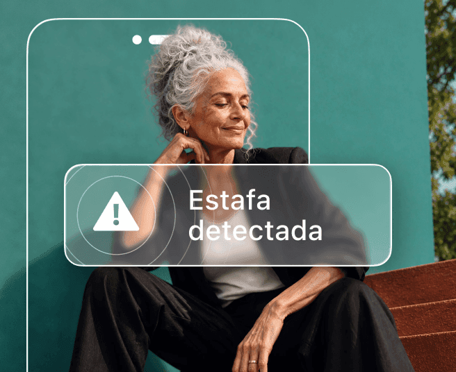 Una mujer con pelo canoso está sentada contra una pared de color verde azulado, parcialmente oculta por la silueta de un teléfono que muestra una alerta de «Se ha detectado una estafa».