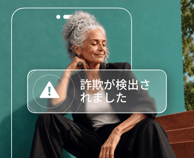 ティール色の壁を背にして座っている白髪の女性。「詐欺が検出されました」というアラートを表示するスマートフォンの輪郭によって、部分的に覆われています。