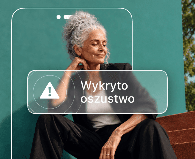 Kobieta z siwymi włosami siedzi na tle zielononiebieskiej ściany, częściowo zasłonięta zarysem telefonu, na którym wyświetla się alert o wykryciu oszustwa.