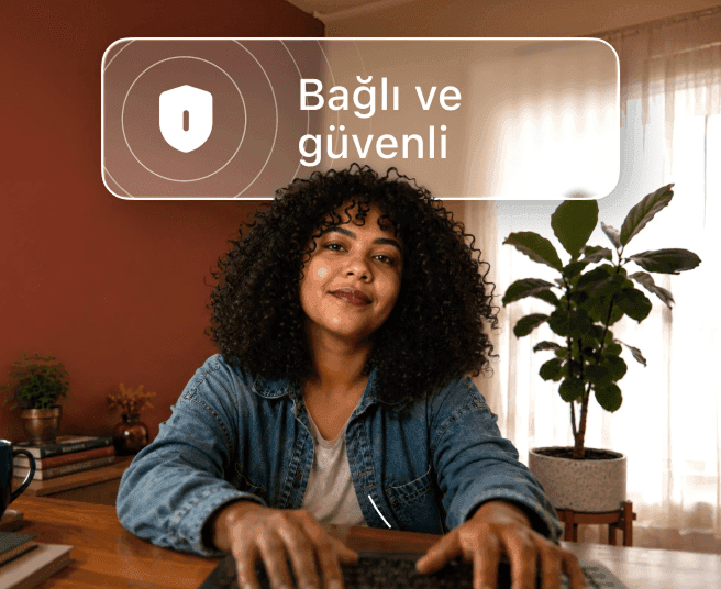 Kıvırcık saçlı bir kişi masada dizüstü bilgisayarda yazı yazıyor ve üzerinde 'Bağlı ve güvende' yazan bir konuşma balonu var.