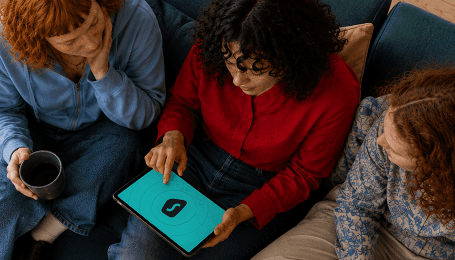 Trois personnes assises sur un canapé regardent l’écran d’une tablette sur lequel est affiché le logo de Surfshark.
