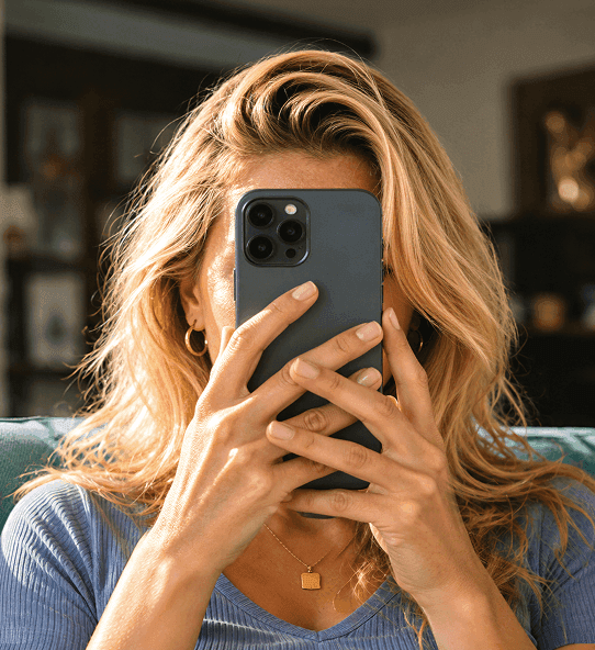 Une femme blonde assise sur un canapé tient un téléphone devant son visage.