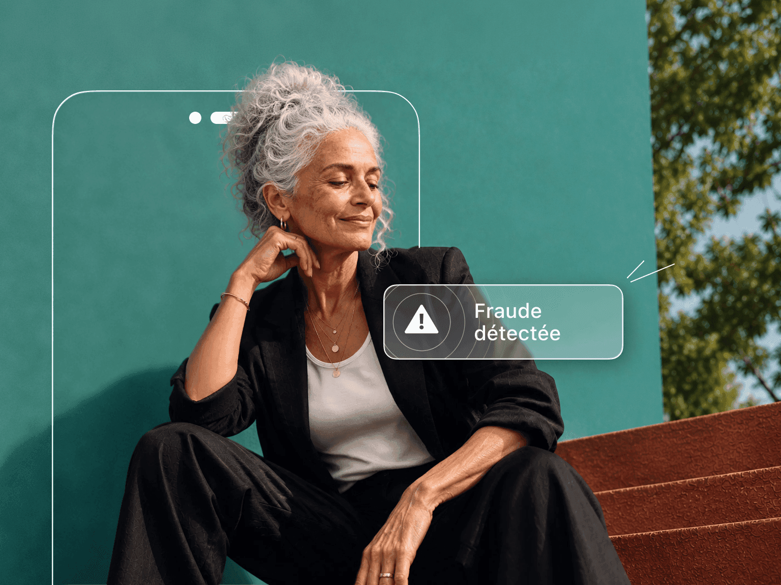 Une femme aux cheveux gris, assise adossée à un mur bleu sarcelle, est partiellement masquée par la silhouette d’un téléphone sur lequel s’affiche une alerte « Fraude détectée ».