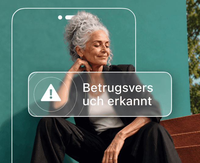 Eine Frau mit grauem Haar sitzt vor einer blaugrünen Wand, teilweise verdeckt durch den Umriss eines Telefons, das einen „Betrug erkannt“-Alert anzeigt.