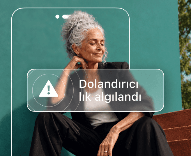 Gri saçlı bir kadın deniz mavisi bir duvarın önünde oturuyor ve Dolandırıcılık tespit edildi Alert gösteren bir telefon taslağı tarafından kısmen gizleniyor.