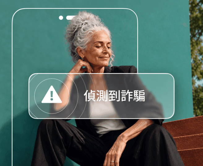 一名灰髮女子坐在藍綠色牆面前,部分身體被手機輪廓遮住,手機畫面顯示「偵測到詐騙」警示。
