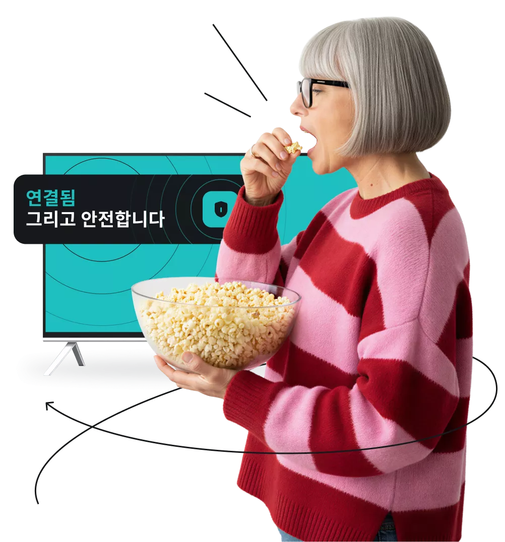 분홍색 및 붉은색 줄무늬 스웨터를 입은 한 여성이 TV 앞에서 팝콘을 먹고 있습니다. TV 화면에는 ‘연결되어 있으며 안전합니다’라는 문구가 표시되어 있습니다.
